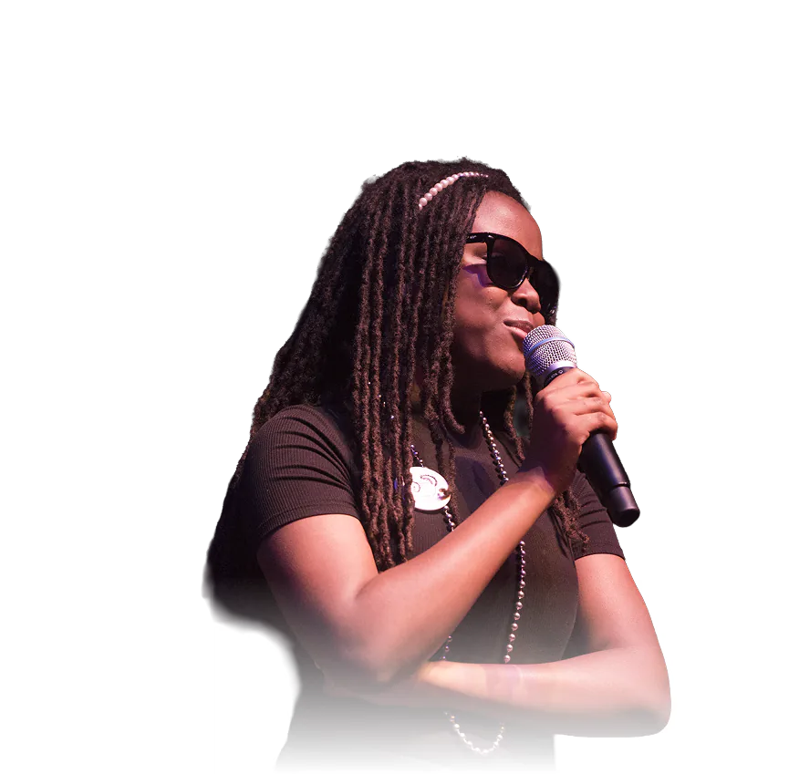 Woman with long locs and sunglasses singing into a microphone while wearing a black T-shirt.